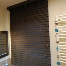 Ciemnobrązowe żaluzje poziome z drewna w łazience, widoczne sznurki sterujące, fragment lustra i dekoracyjny grzejnik drabinkowy