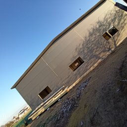 USŁUGI REMONTOWO-BUDOWLANE DOMINIK NOWAK - Nowoczesny budynek przemysłowy z szarej blachy trapezowej, widok z dołu, z widocznymi otworami okiennymi i cieniem drzewa na elewacji, na pierwszym planie leżące panele.