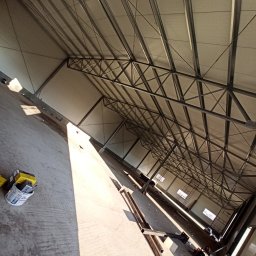 USŁUGI REMONTOWO-BUDOWLANE DOMINIK NOWAK - Wnętrze hali w trakcie budowy z widoczną konstrukcją dachu, stalowymi elementami nośnymi i panelami dachowymi. Na podłodze widoczne materiały budowlane i narzędzia.