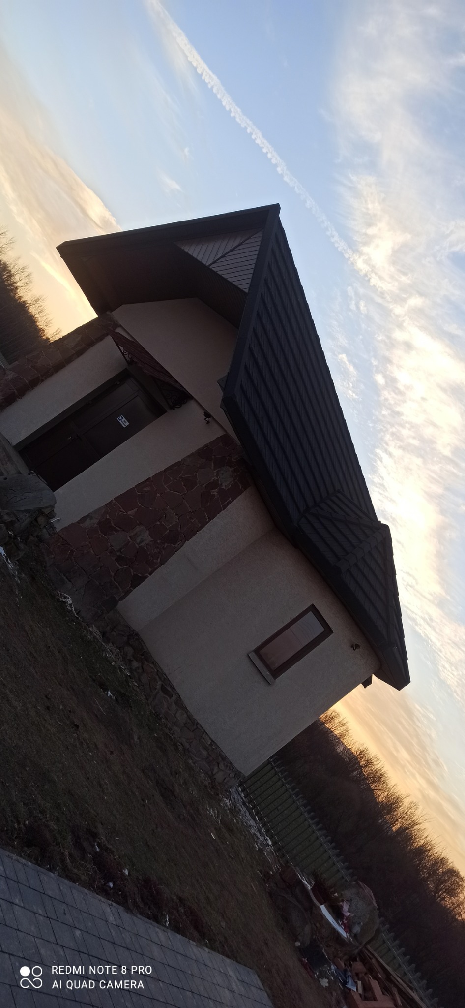 Nowoczesny budynek z ciemnym, skośnym dachem z blachy trapezowej, wykończony kamieniem elewacyjnym, sfotografowany pod kątem z widocznym śladem kondensacyjnym na niebie.