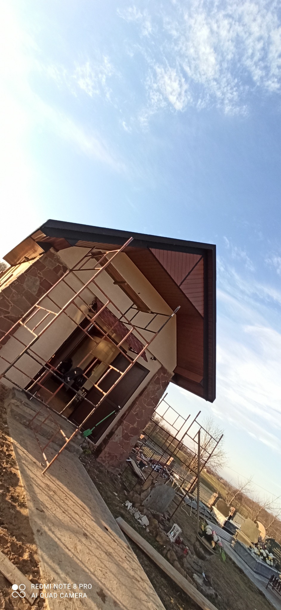 Nisko kątowe ujęcie domu z nowym, ciemnym dachem i kamienną elewacją w Waśniowie, częściowo zasłoniętego przez metalowe rusztowanie, na tle błękitnego nieba z delikatnymi chmurami.