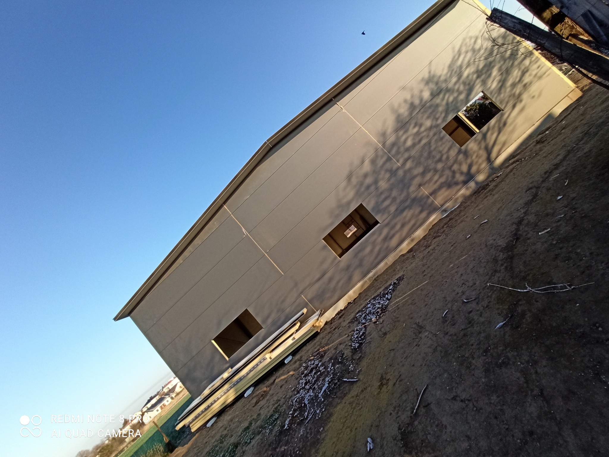 Nowoczesny budynek przemysłowy z szarej blachy trapezowej, widok z dołu, z widocznymi otworami okiennymi i cieniem drzewa na elewacji, na pierwszym planie leżące panele.