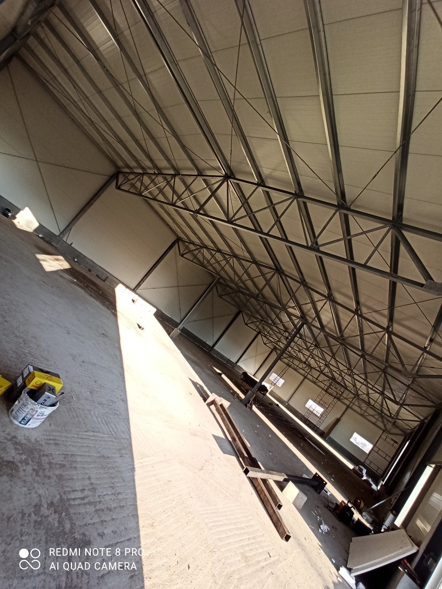 Wnętrze hali w trakcie budowy z widoczną konstrukcją dachu, stalowymi elementami nośnymi i panelami dachowymi. Na podłodze widoczne materiały budowlane i narzędzia.