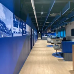 Długi korytarz w barwach klubu sportowego z dekoracyjnymi panelami ściennymi z motywami piłkarskimi, nowoczesne oświetlenie sufitowe, stoliki i krzesła w strefie gastronomicznej.