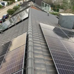 Widok z g&oacute;ry na dach pokryty dach&oacute;wką z zainstalowanymi panelami fotowoltaicznymi, odbijającymi niebo i otoczenie. W tle widoczne budynki mieszkalne i drzewa.