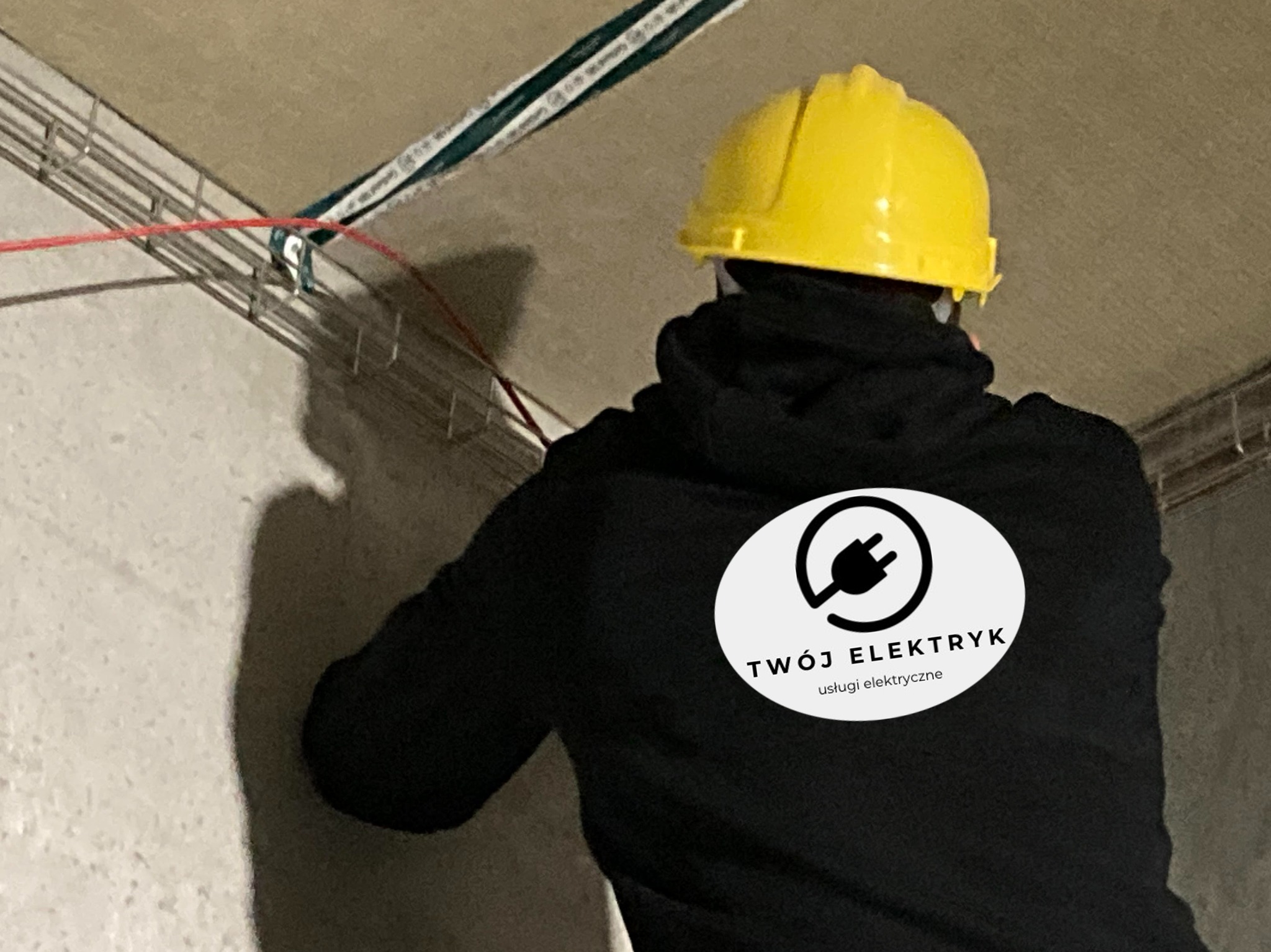 Elektryk w czarnej bluzie z logo firmy na plecach i żółtym kasku, montuje korytka kablowe pod sufitem w surowym pomieszczeniu.