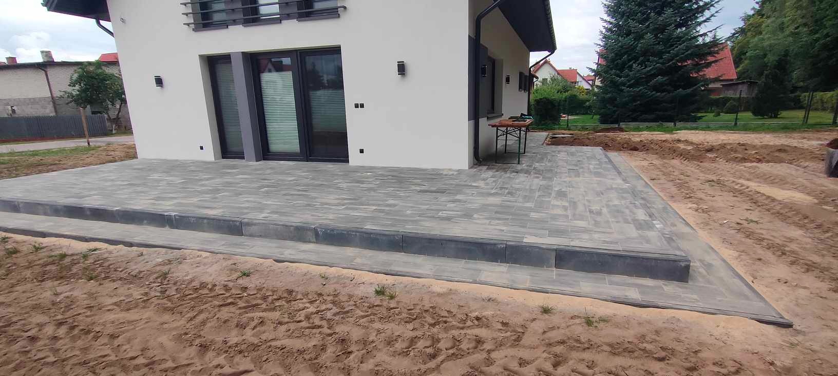 Nowoczesny taras z szarej kostki brukowej ułożony przed białym domem z czarnymi ramami okiennymi; widoczne ślady prac ziemnych wokół tarasu.