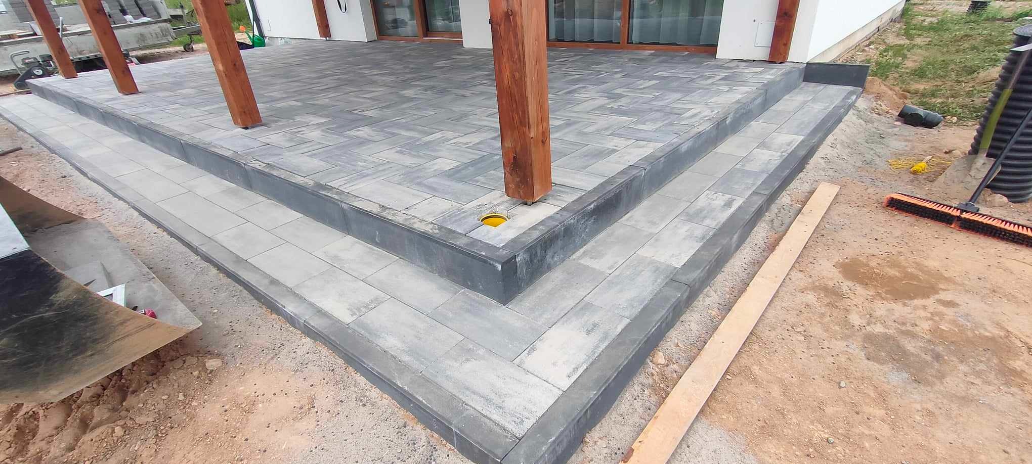 Nowo ułożony taras z szarej kostki brukowej ułożonej we wzór jodełki, z drewnianymi słupami podtrzymującymi zadaszenie i żółtą rurą drenażową w rogu.