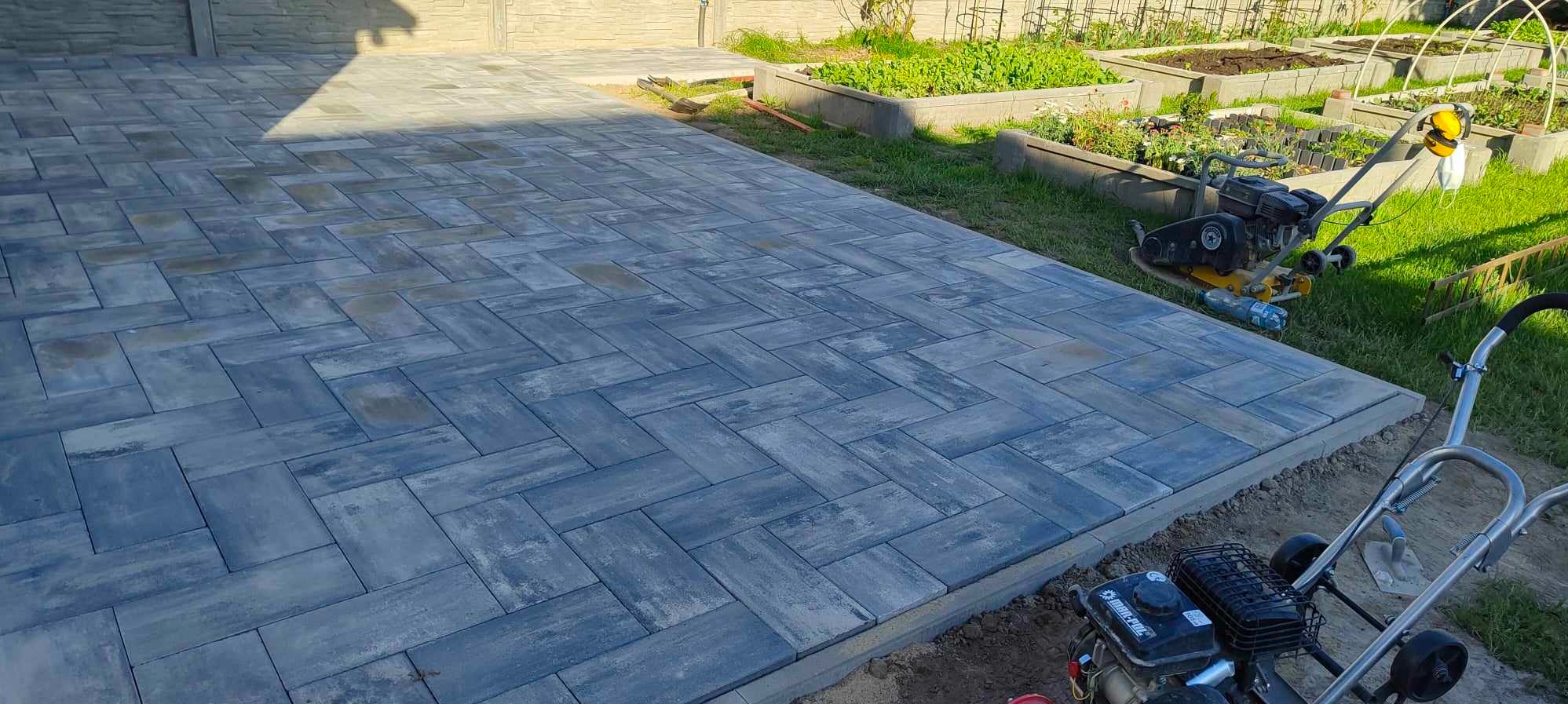 Ułożona, szara kostka brukowa w jodełkę na posesji, widoczne zagony z roślinami i sprzęt budowlany