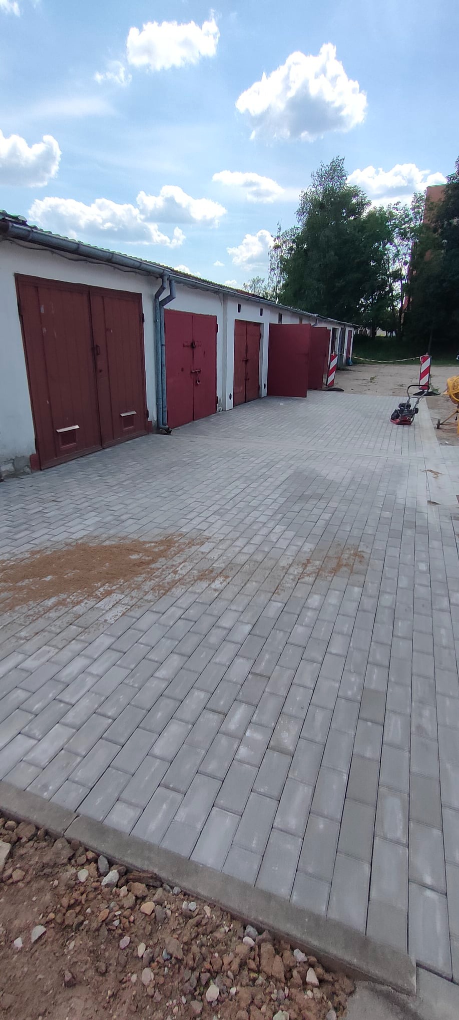 Świeżo ułożona, szara kostka brukowa przed szeregiem garaży z czerwonymi drzwiami, widoczne narzędzia i piasek na powierzchni, błękitne niebo z chmurami w tle.
