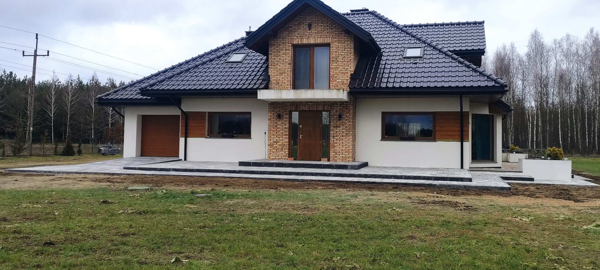 Nowoczesny dom z ciemną dachówką i jasną elewacją. Ułożona kostka brukowa tworzy chodnik i podjazd. Tło stanowią drzewa bez liści.
