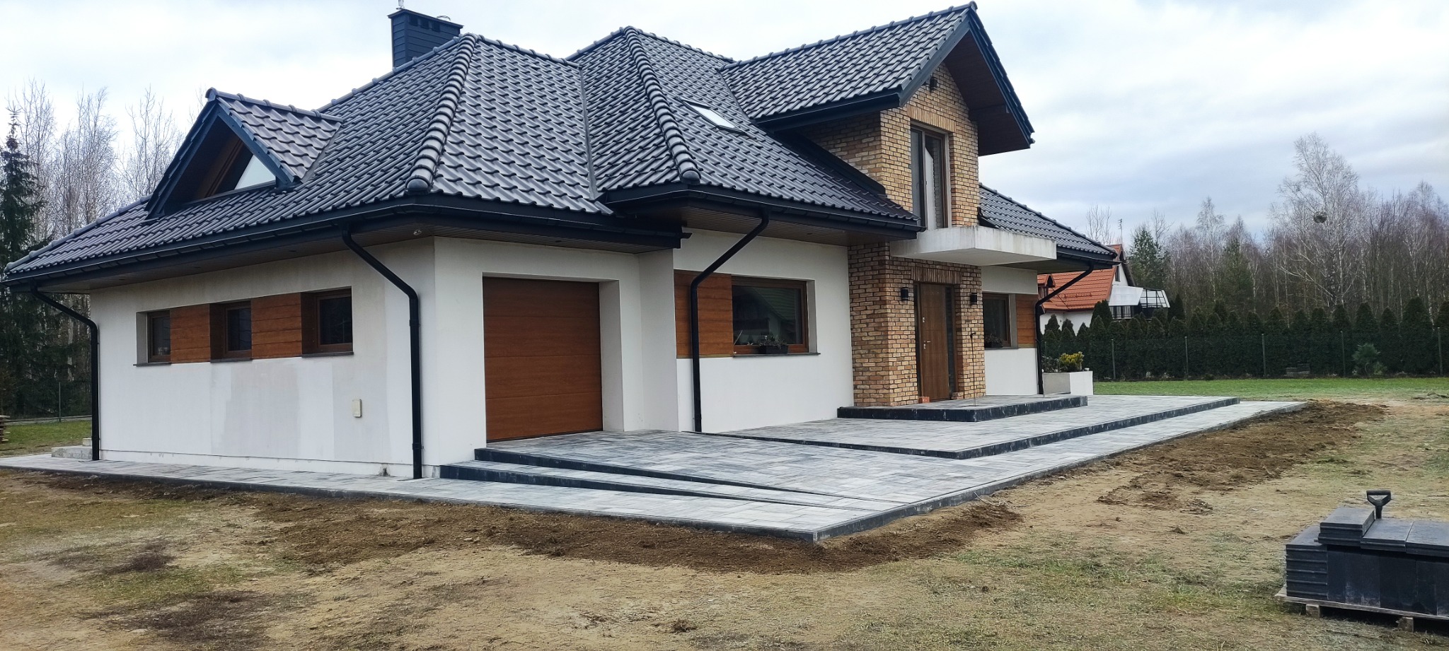 Nowoczesny dom z szarej kostki brukowej na podjeździe i schodach wejściowych. Widoczne niedokończone prace ziemne wokół posesji i paleta z kostką. Pochmurne niebo.