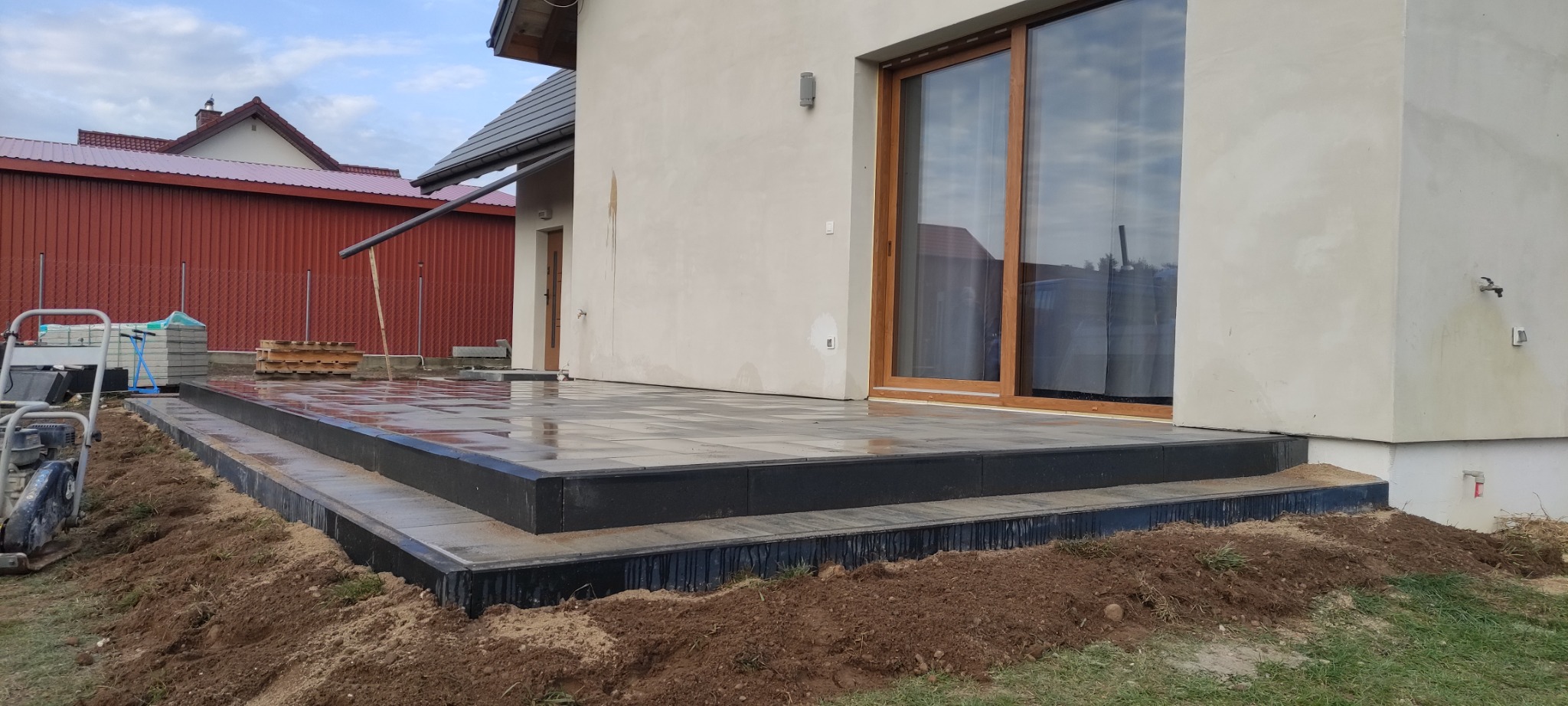 Nowoczesny taras z kostki brukowej w odcieniach szarości, z czarnymi obrzeżami, przylegający do jasnego budynku z dużymi oknami przesuwnymi. Widoczna ubijarka i kostka.