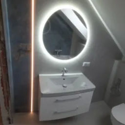 Wykończona łazienka w Głownie z umywalką na szafce, okrągłym lustrem z podświetleniem LED i szarymi płytkami. Widoczny fragment ściany w stanie surowym.