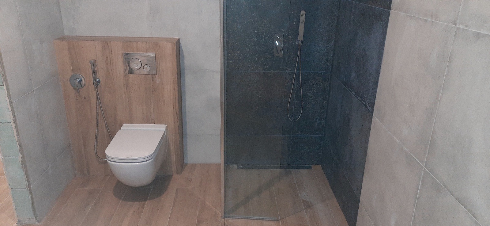 Nowoczesna łazienka z kabiną prysznicową typu walk-in wyłożoną ciemnymi płytkami, podwieszaną toaletą z drewnianą obudową i bidetką oraz podłogą imitującą drewno.