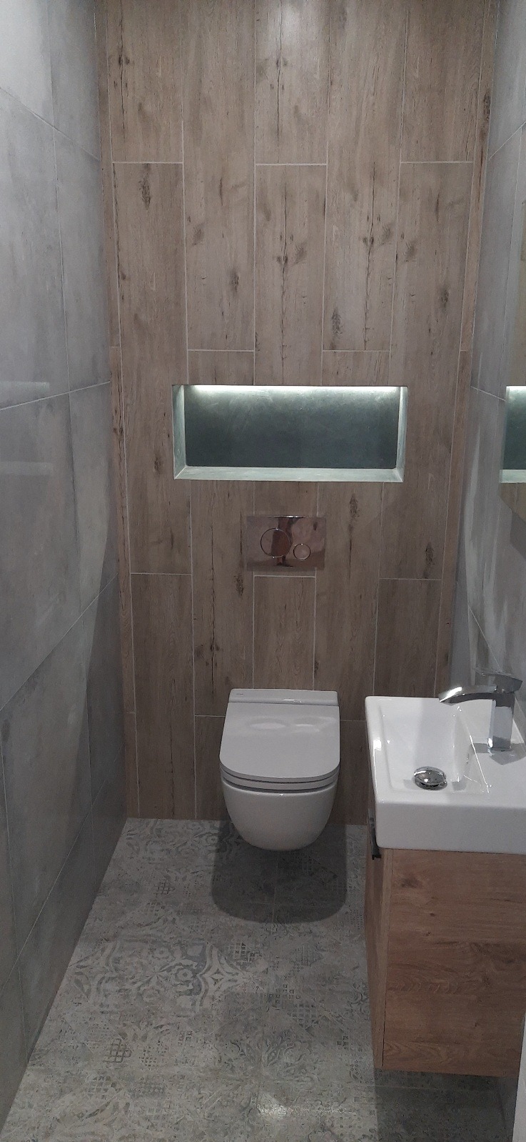 Nowoczesna łazienka po remoncie: płytki imitujące drewno na ścianach, podświetlana wnęka, podłoga w dekoracyjne wzory, wisząca toaleta i umywalka z drewnianą szafką.