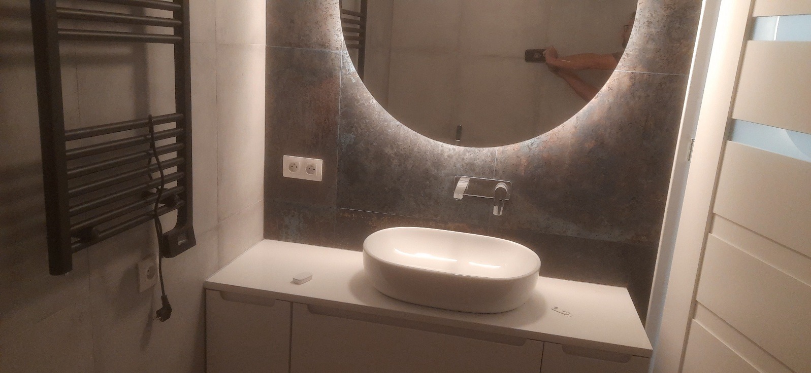 Nowoczesna łazienka z okrągłym lustrem podświetlanym LED, białą umywalką nablatową i grzejnikiem drabinkowym w ciemnym kolorze. Minimalistyczny design i ciepłe oświetlenie.