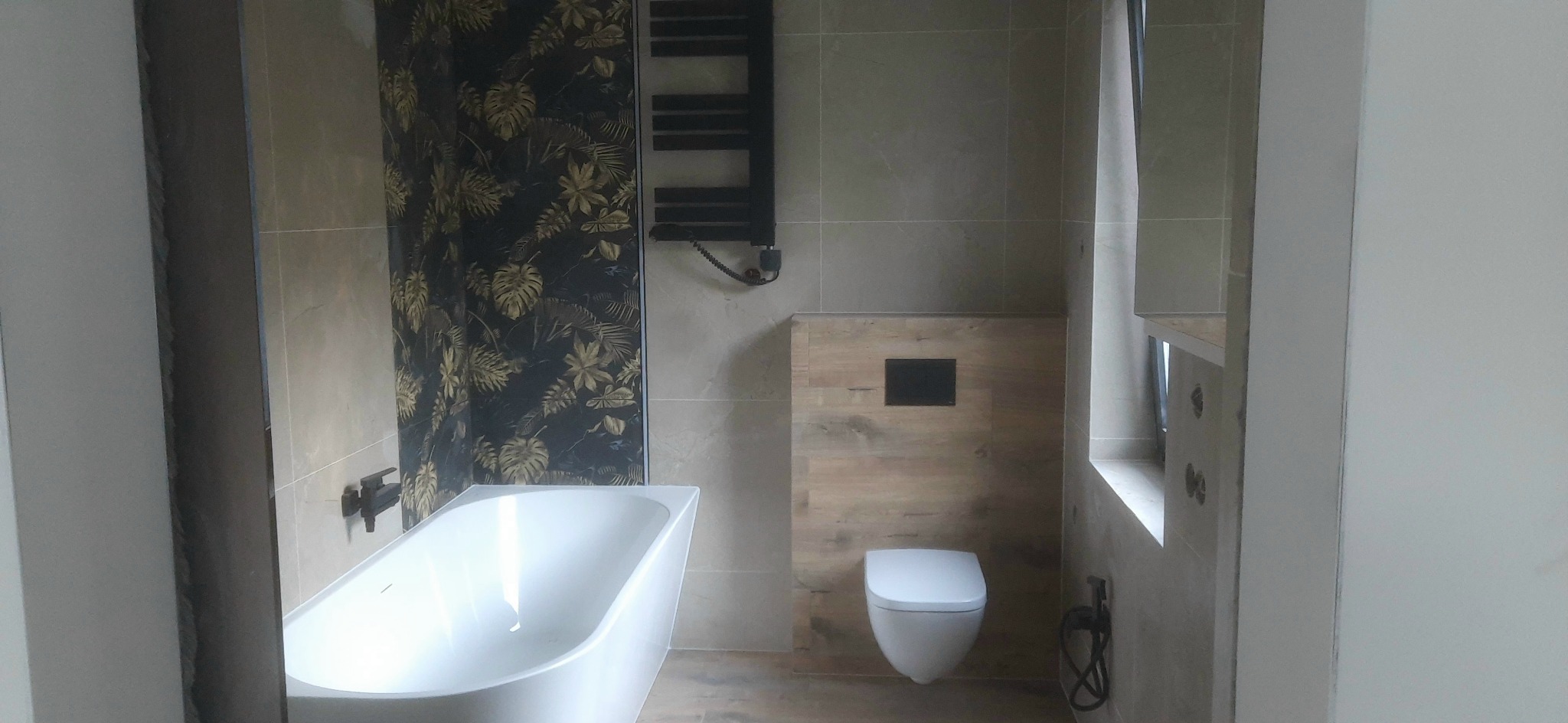 Elegancka łazienka po remoncie: wanna z armaturą, toaleta podtynkowa z drewnianym panelem, grzejnik drabinkowy na tle płytek z motywem roślinnym.