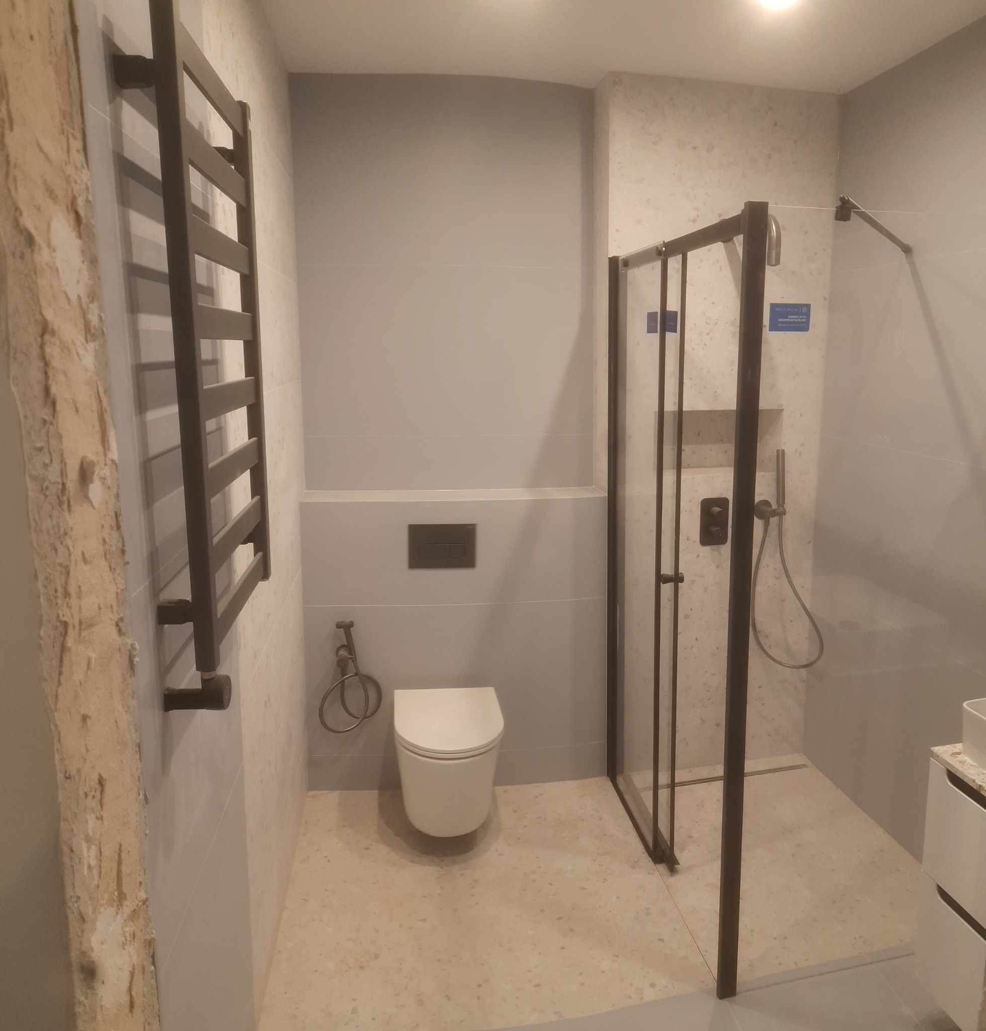 Nowoczesna łazienka po remoncie z kabiną prysznicową z czarnymi ramami, grzejnikiem drabinkowym i podtynkową toaletą. Jasne płytki i minimalistyczny design.