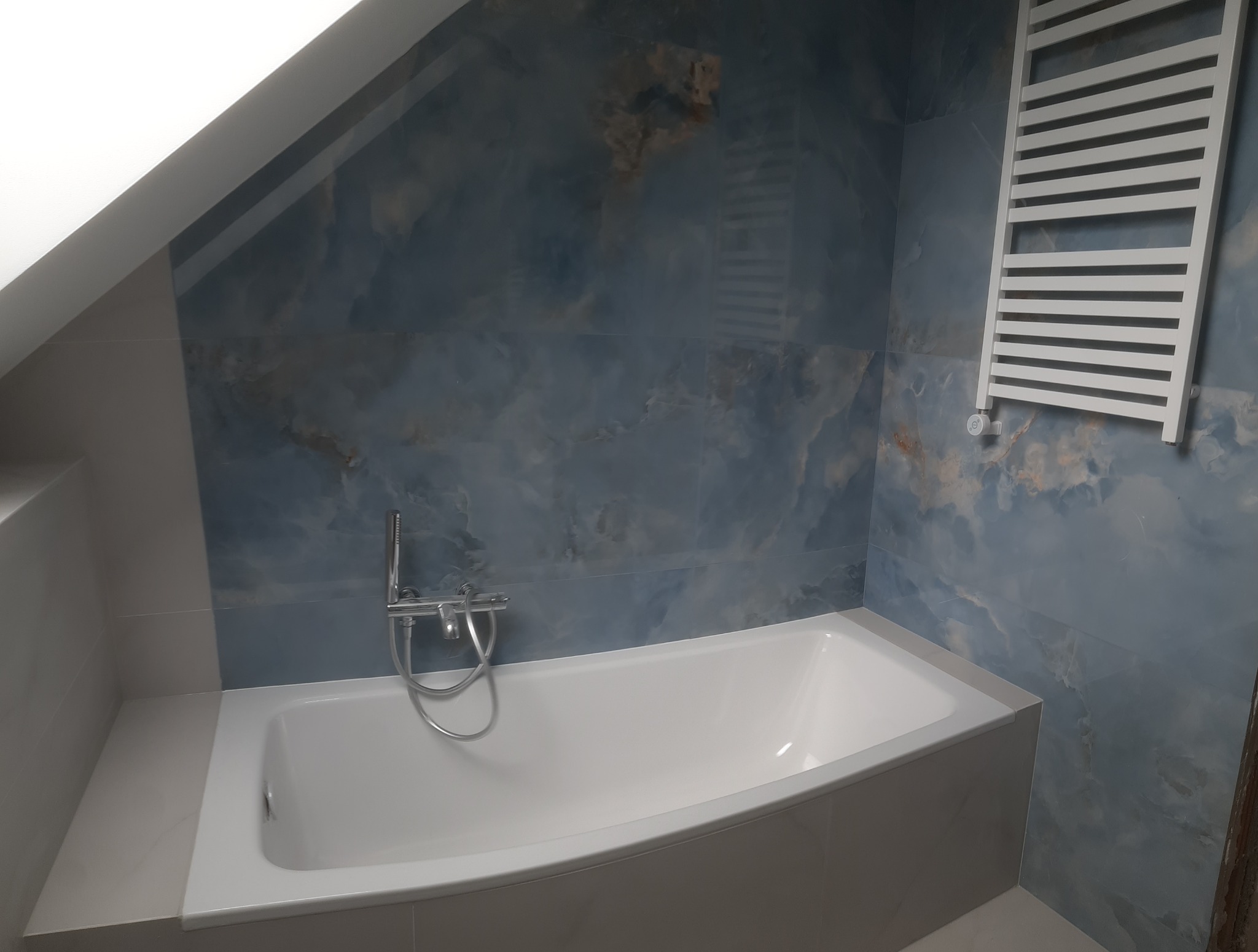 Wanna w łazience na poddaszu z błękitnymi płytkami imitującymi marmur. Grzejnik drabinkowy na ścianie. Bateria wannowa chromowana. Całość utrzymana w chłodnej tonacji.