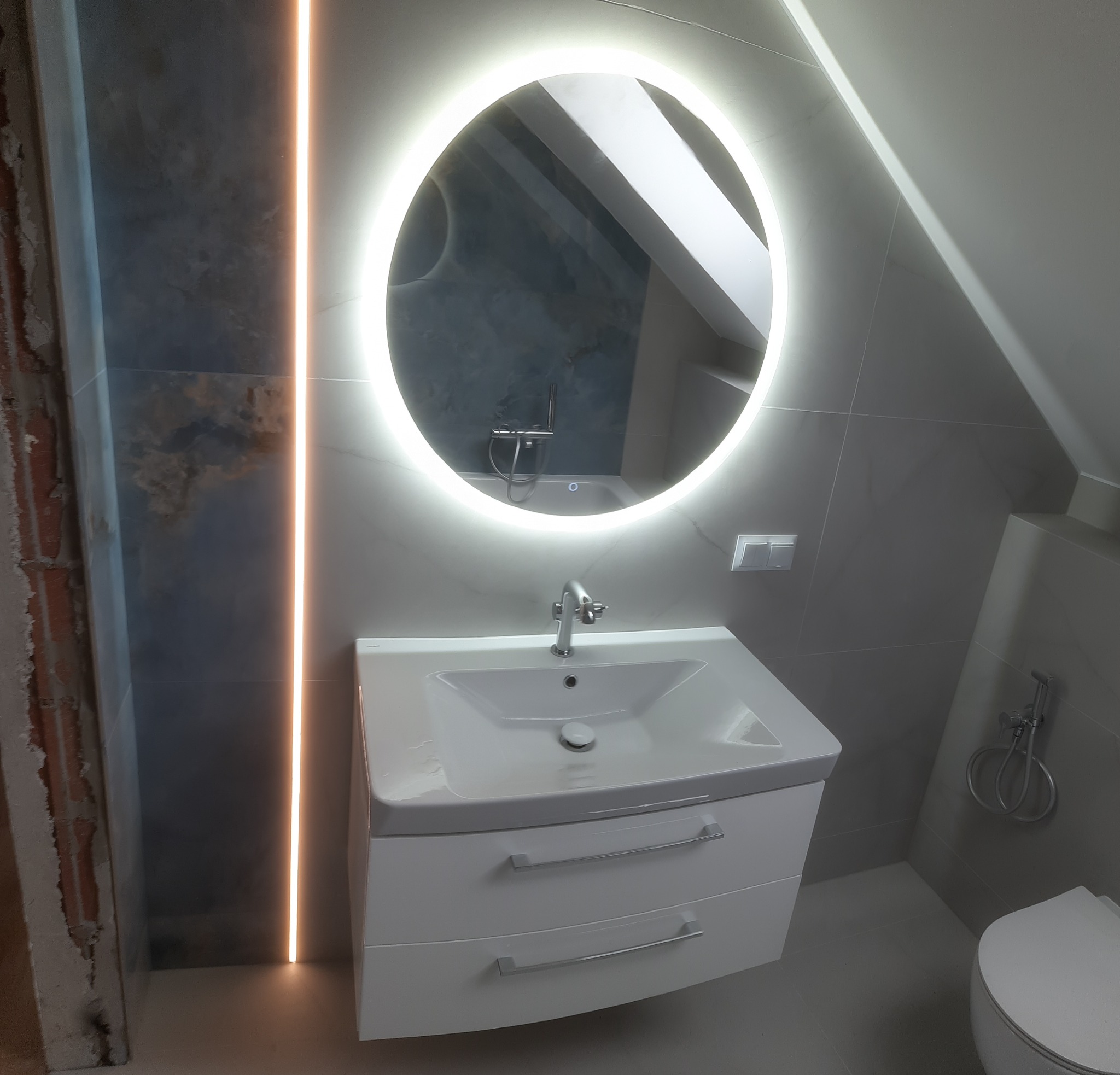 Wykończona łazienka w Głownie z umywalką na szafce, okrągłym lustrem z podświetleniem LED i szarymi płytkami. Widoczny fragment ściany w stanie surowym.