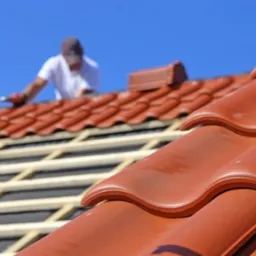 Układanie czerwonej dach&oacute;wki ceramicznej na drewnianej konstrukcji dachu, widoczny pracownik w tle, błękitne niebo.