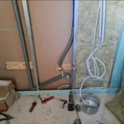 Wnętrze w trakcie prac instalacyjnych: widoczne podejścia kanalizacyjne i wodne z rur PCV, przewody elektryczne w karbowanych peszlach, ściany w trakcie zabudowy płytami gipsowo-kartonowymi...