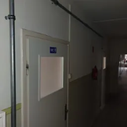 Szara rura instalacji przeciwpożarowej, zamocowana do ściany korytarza z drzwiami do toalety i gaśnicą w schronisku.