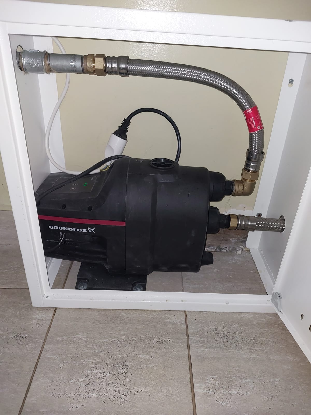 Czarna pompa hydroforowa Grundfos z podłączonymi przewodami elastycznymi w metalowym oplocie, zamontowana w białej szafce instalacyjnej na płytkach podłogowych.