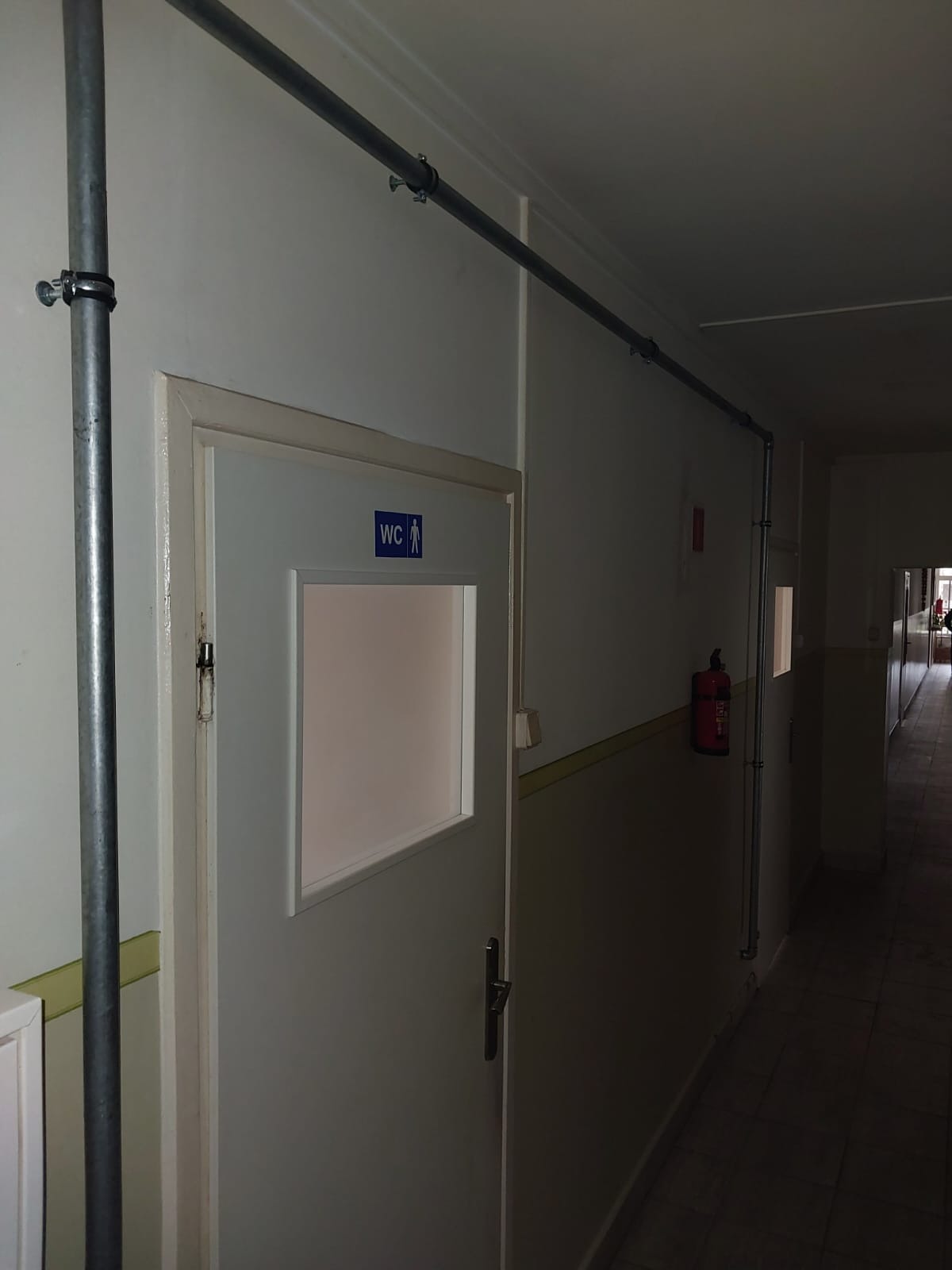 Szara rura instalacji przeciwpożarowej, zamocowana do ściany korytarza z drzwiami do toalety i gaśnicą w schronisku.