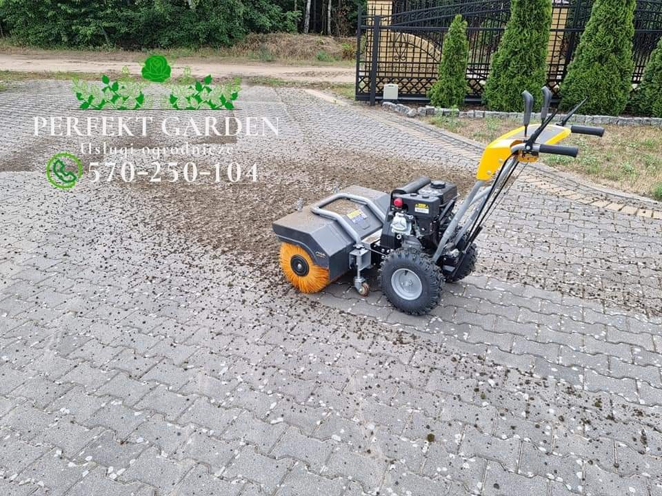 Szczotka mechaniczna do czyszczenia kostki brukowej z resztek roślinnych, widoczny efekt czyszczenia na szarej kostce, w tle drzewa i metalowe ogrodzenie.