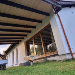 Biała elewacja domu z drewnianą konstrukcją zadaszenia tarasu, widoczne okna z drewnianymi ramami, brązowe rynny i wózek z drewnem na trawniku.