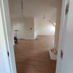 Widok z perspektywy otwartych drzwi na wykończone wnętrze z drewnianą podłogą, surowe kable zwisające z sufitu i sprzęt budowlany w tle.