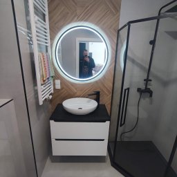 F.U. COMPLEX CONSTRUCTION WOJCIECH WOŹNIAK - Nowoczesna łazienka z umywalką nablatową, podświetlanym lustrem, grzejnikiem drabinkowym i kabiną prysznicową z czarnymi elementami, wykończona płytkami imitującymi drewno.