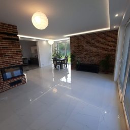 F.U. COMPLEX CONSTRUCTION WOJCIECH WOŹNIAK - Przestronne wnętrze z błyszczącą, białą podłogą z płytek, ceglaną ścianą i kominkiem, oświetlone kulistymi lampami i listwami LED pod sufitem.