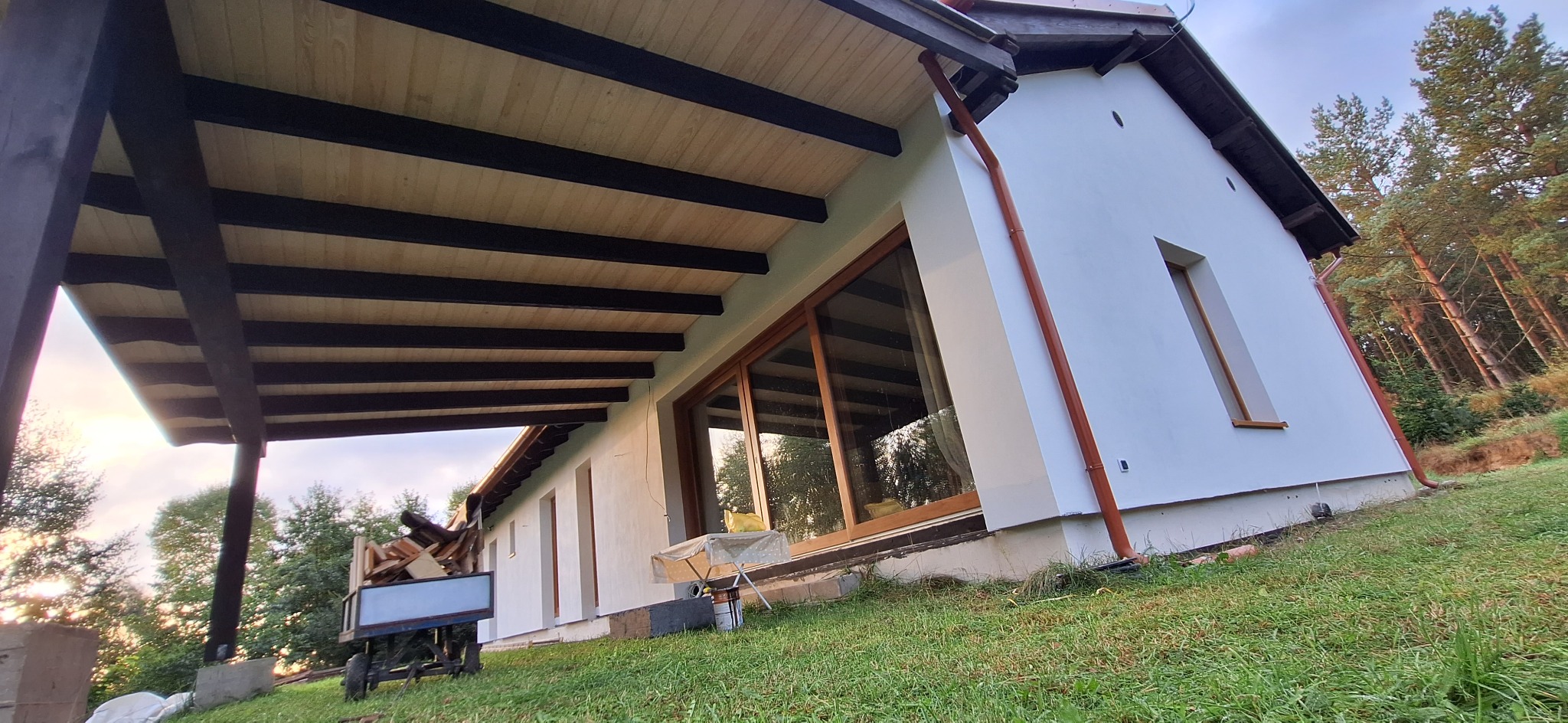Biała elewacja domu z drewnianą konstrukcją zadaszenia tarasu, widoczne okna z drewnianymi ramami, brązowe rynny i wózek z drewnem na trawniku.