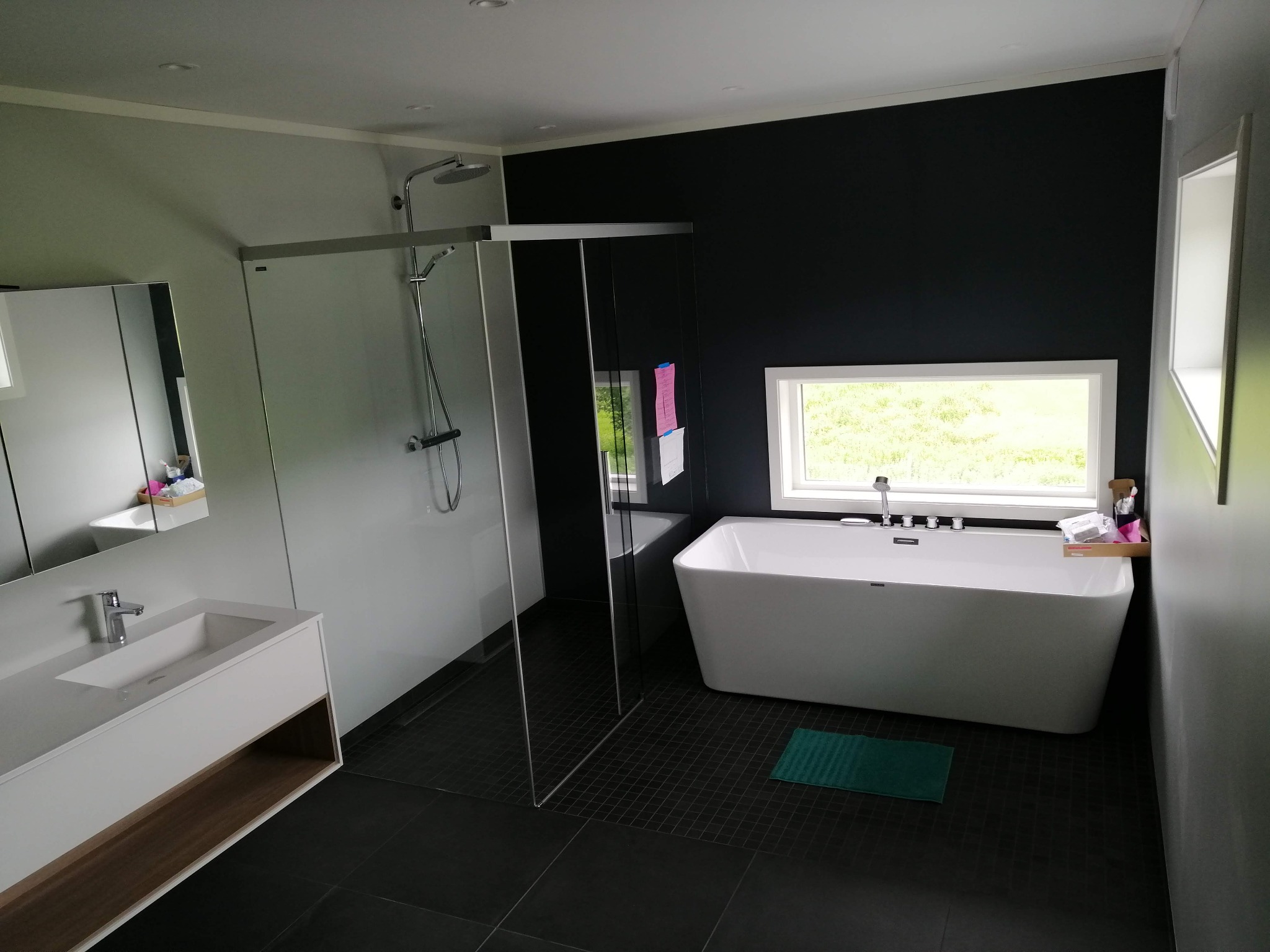 Nowoczesna łazienka z czarną ścianą, białą wanną wolnostojącą, kabiną prysznicową typu walk-in i minimalistyczną umywalką z szafką, wykończona ciemną, kwadratową glazurą podłogową.