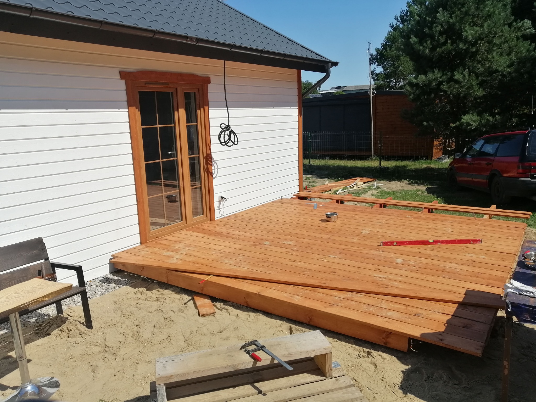 Budowa drewnianego tarasu przy domu z białymi ścianami, widoczny poziomica, zacisk stolarski i wiadro z farbą, piasek wokół tarasu.