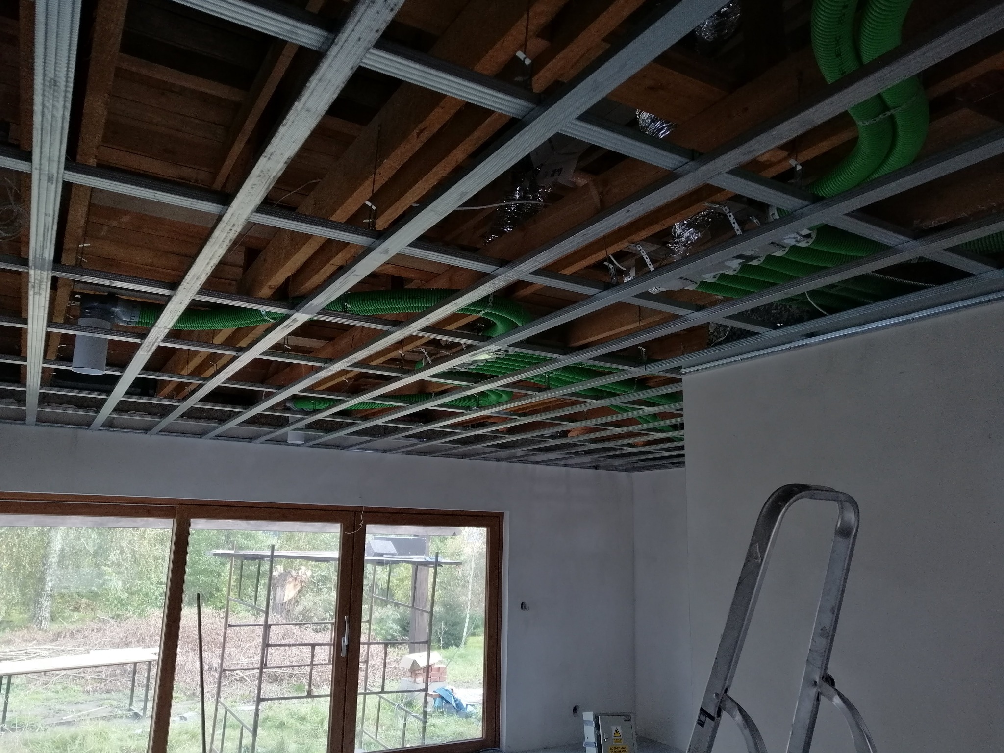 Montaż sufitu podwieszanego z widocznymi instalacjami wentylacyjnymi w Tczewie, widok na konstrukcję z profili stalowych i zielone rury wentylacyjne nad oknem.