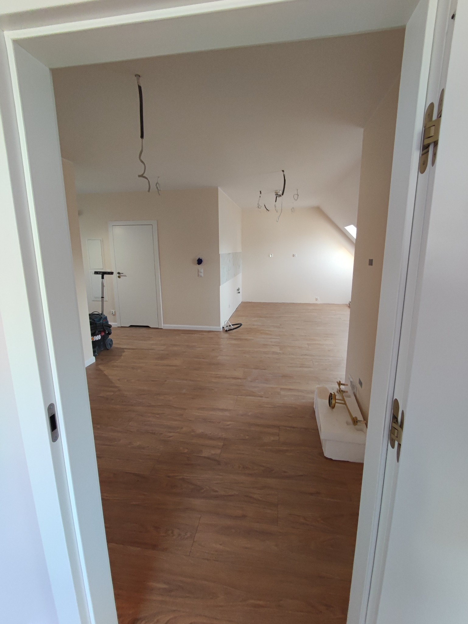 Widok z perspektywy otwartych drzwi na wykończone wnętrze z drewnianą podłogą, surowe kable zwisające z sufitu i sprzęt budowlany w tle.