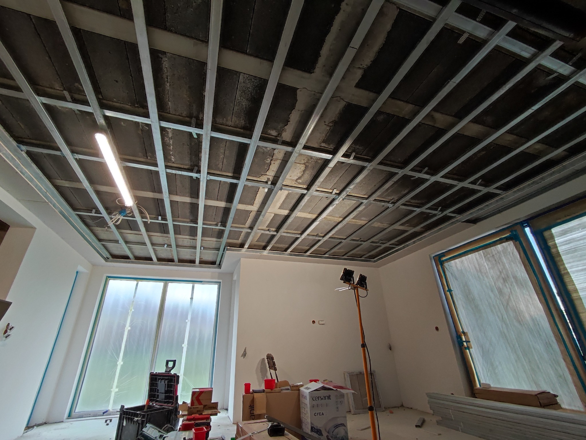 Montaż stelaża sufitu podwieszanego w pomieszczeniu z zabezpieczonymi oknami folią, widoczna lampa robocza i materiały budowlane.