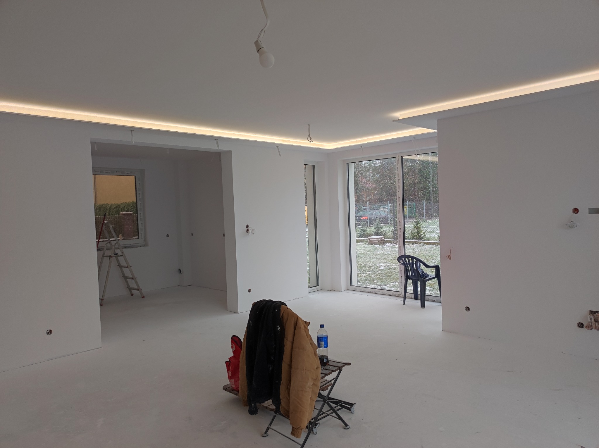Wnętrze w trakcie wykańczania: białe ściany, sufit z oświetleniem LED, drabina oparta o ścianę w sąsiednim pomieszczeniu, krzesło i ubrania robocze na składanym stoliku w centralnej części pokoju.