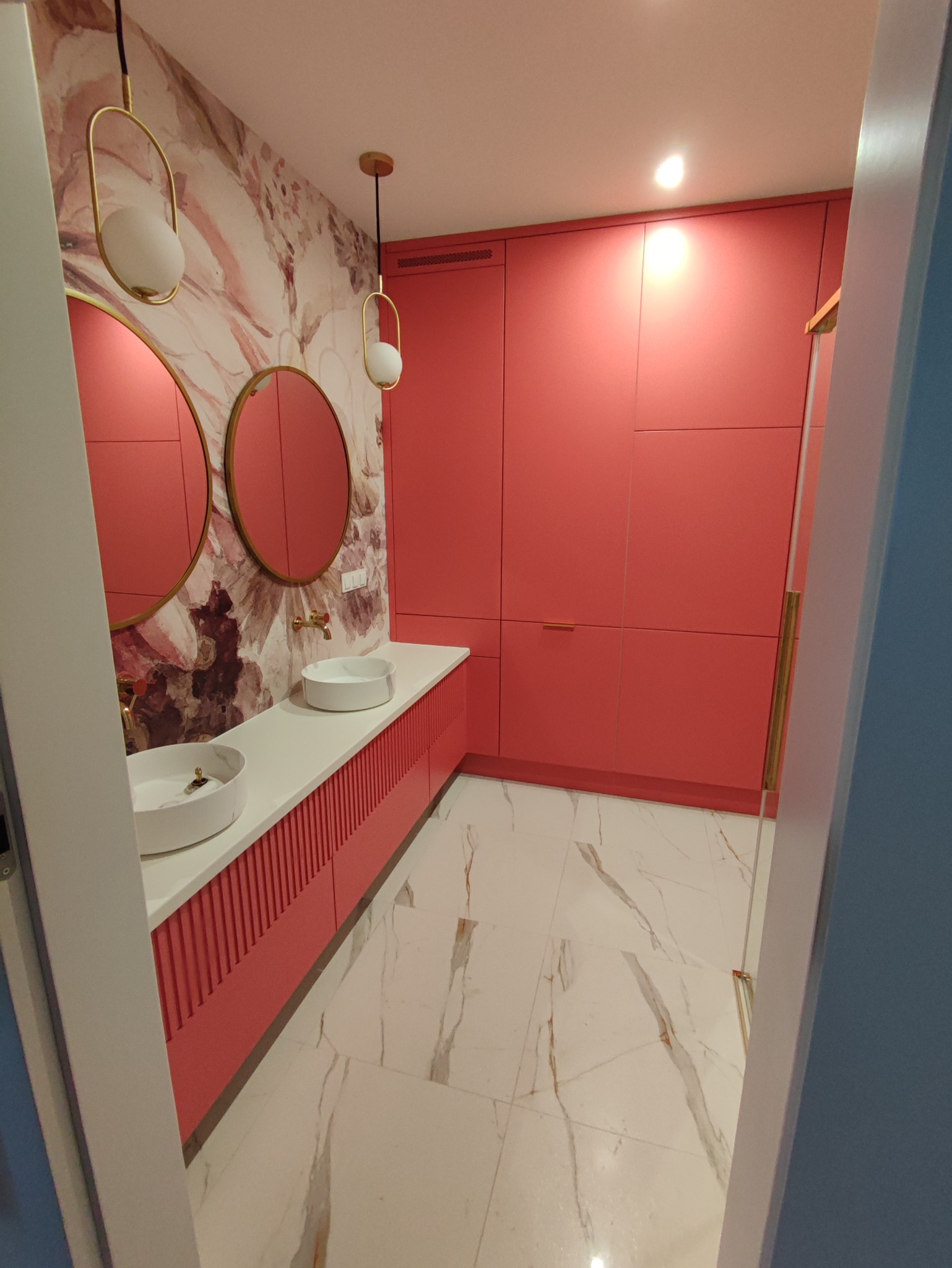 Nowoczesna łazienka z dwoma umywalkami na blacie z ryflowanym frontem w odcieniach koralu, okrągłe lustra z złotymi ramami, ściana w kwiatowy wzór i marmurowa podłoga.