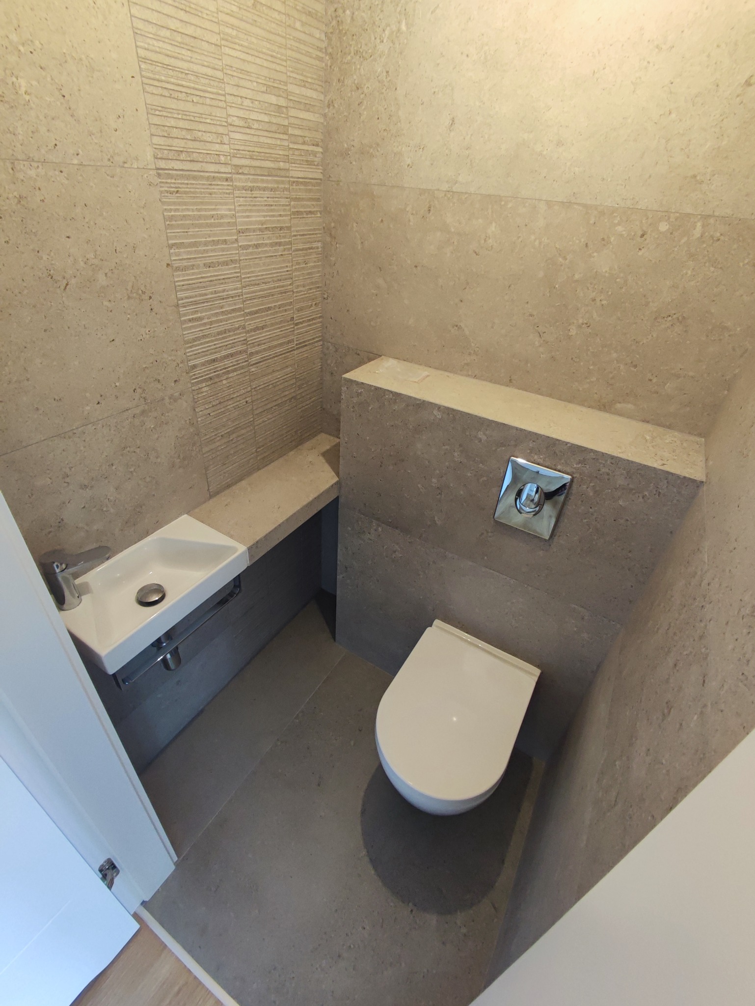 Nowoczesna łazienka w beżowo-szarej tonacji z wiszącą toaletą i umywalką na blacie, ściany wyłożone płytkami o zróżnicowanej fakturze.