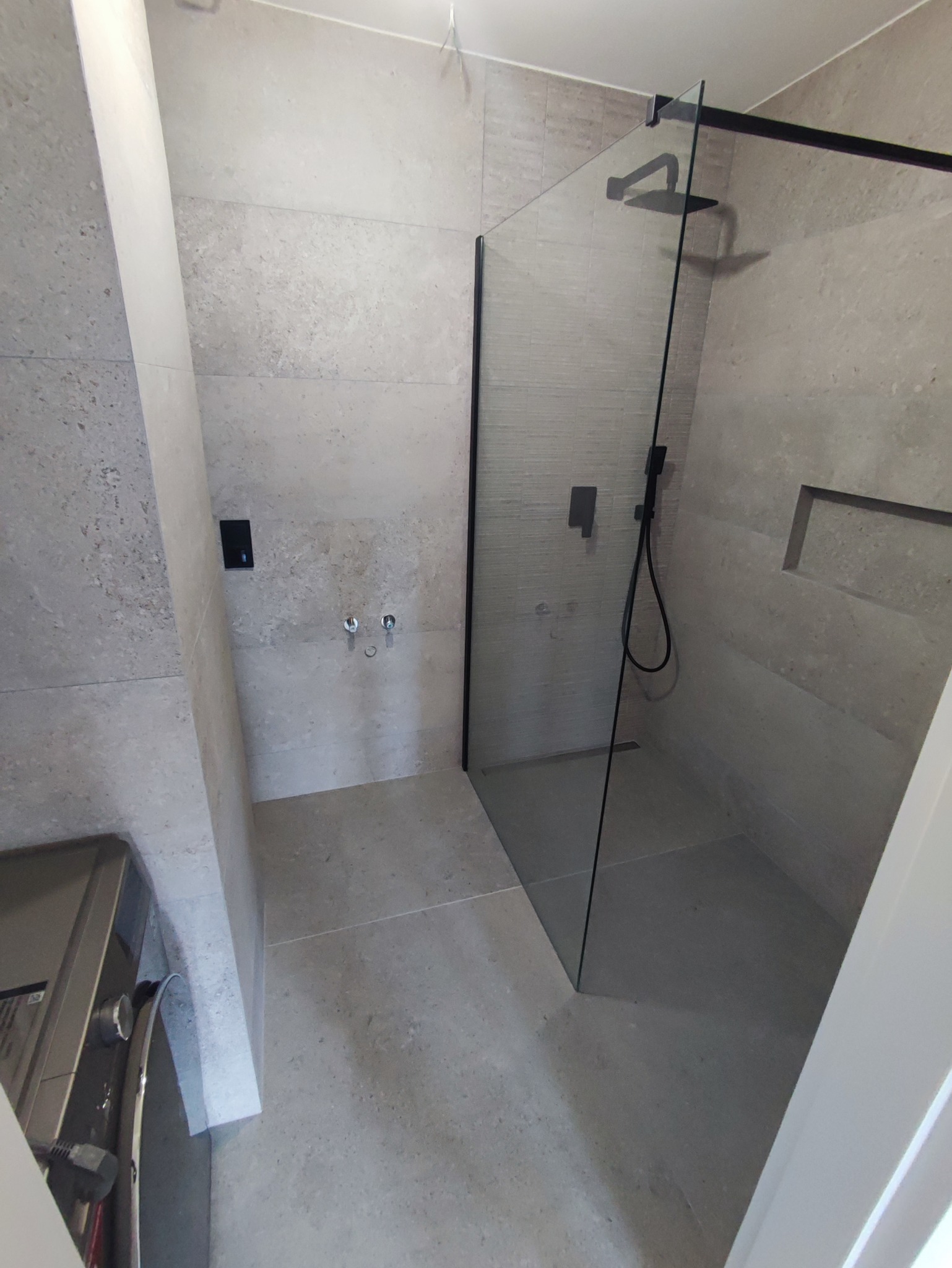 Nowoczesna kabina prysznicowa typu walk-in z czarnymi elementami wykończeniowymi, zamontowana w łazience wyłożonej szarymi płytami imitującymi kamień, z widocznym odpływem liniowym...