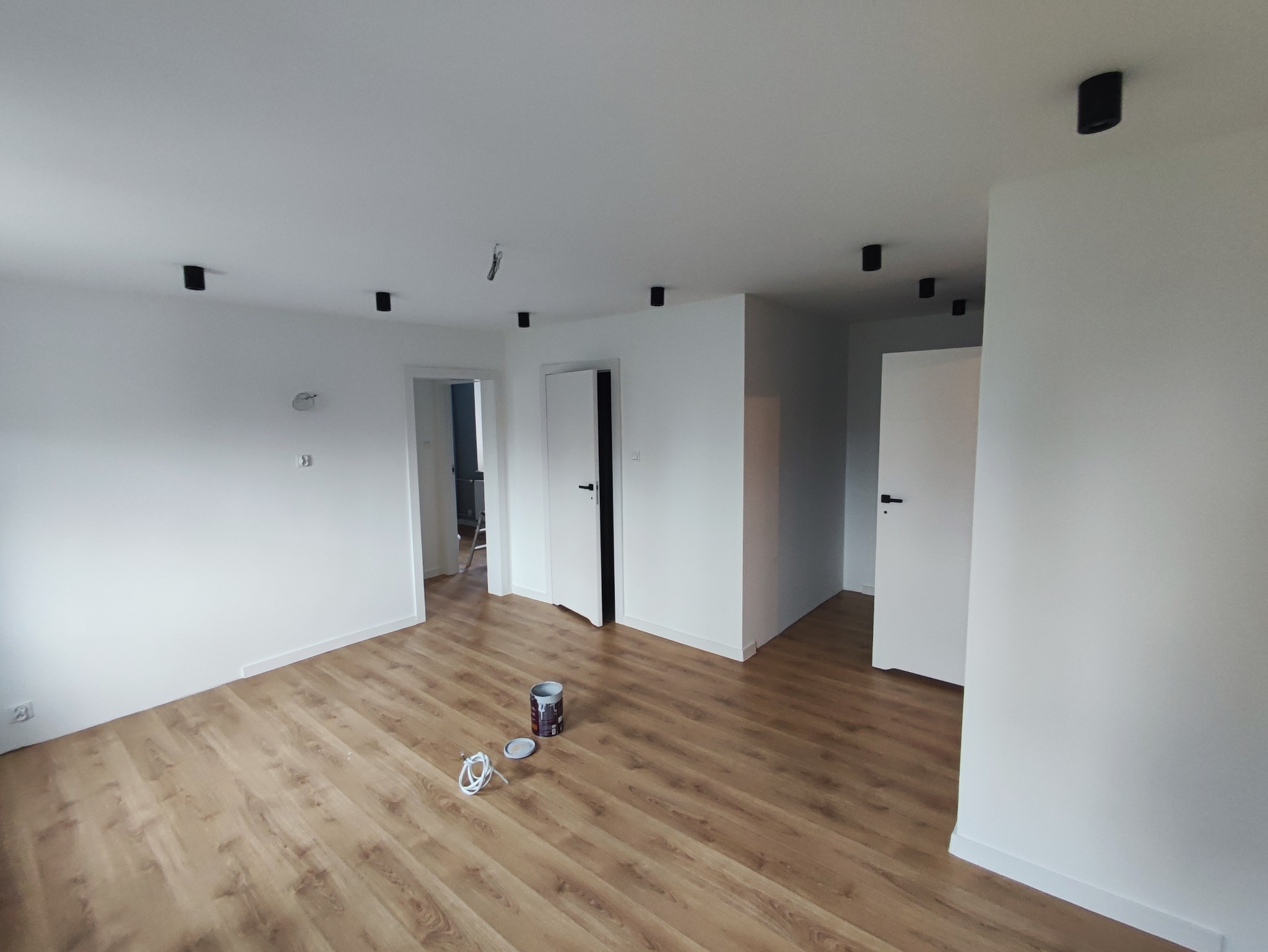 Pomalowane na biało wnętrze mieszkania z panelami podłogowymi, widoczne drzwi wewnętrzne, czarne reflektory sufitowe i puszka farby na podłodze.