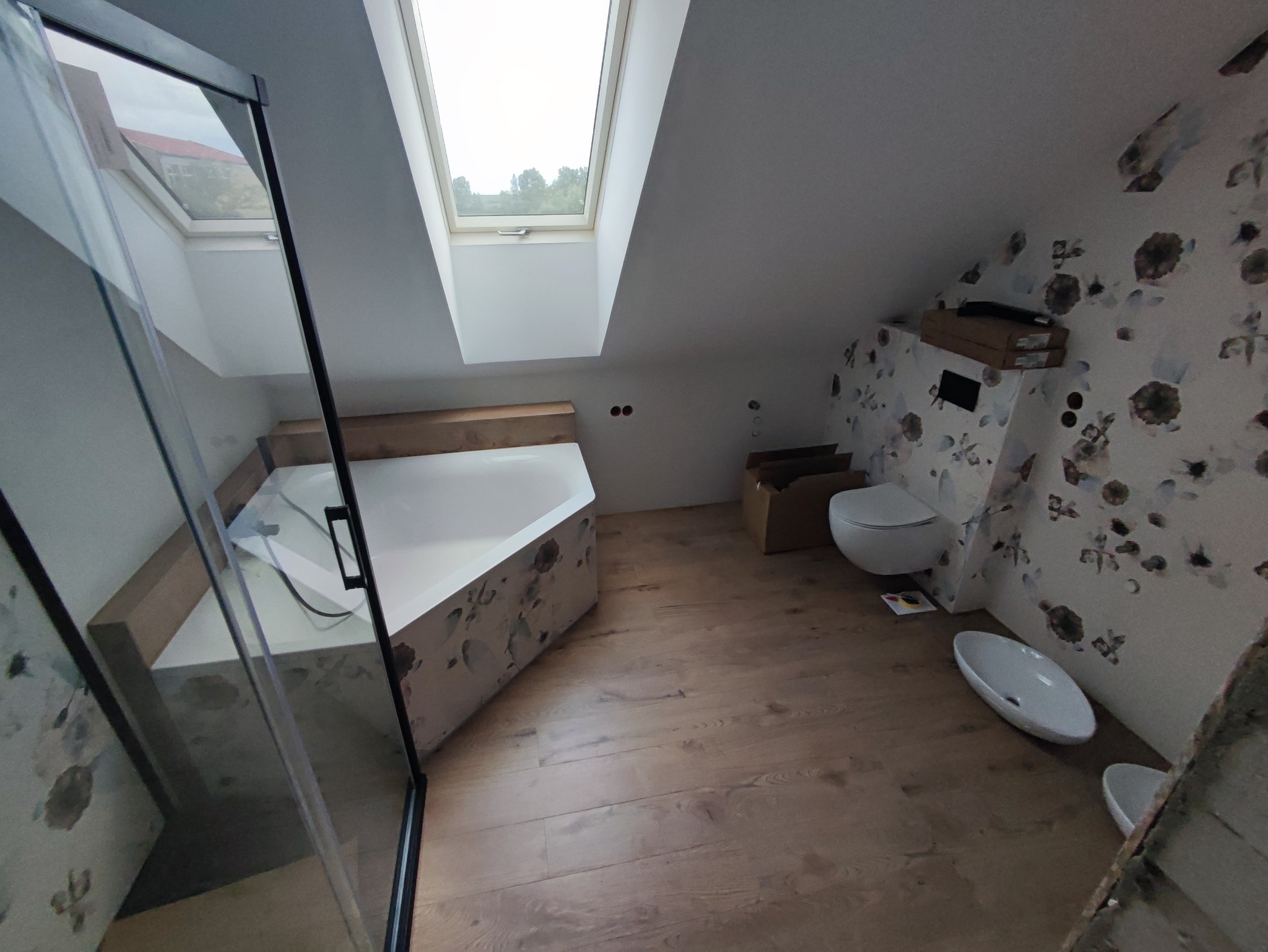 Wykończona łazienka na poddaszu z oknem dachowym, kabiną prysznicową z czarnymi ramami, toaletą podwieszaną, umywalką nablatową i drewnianą podłogą; na ścianie tapeta w kwiaty.