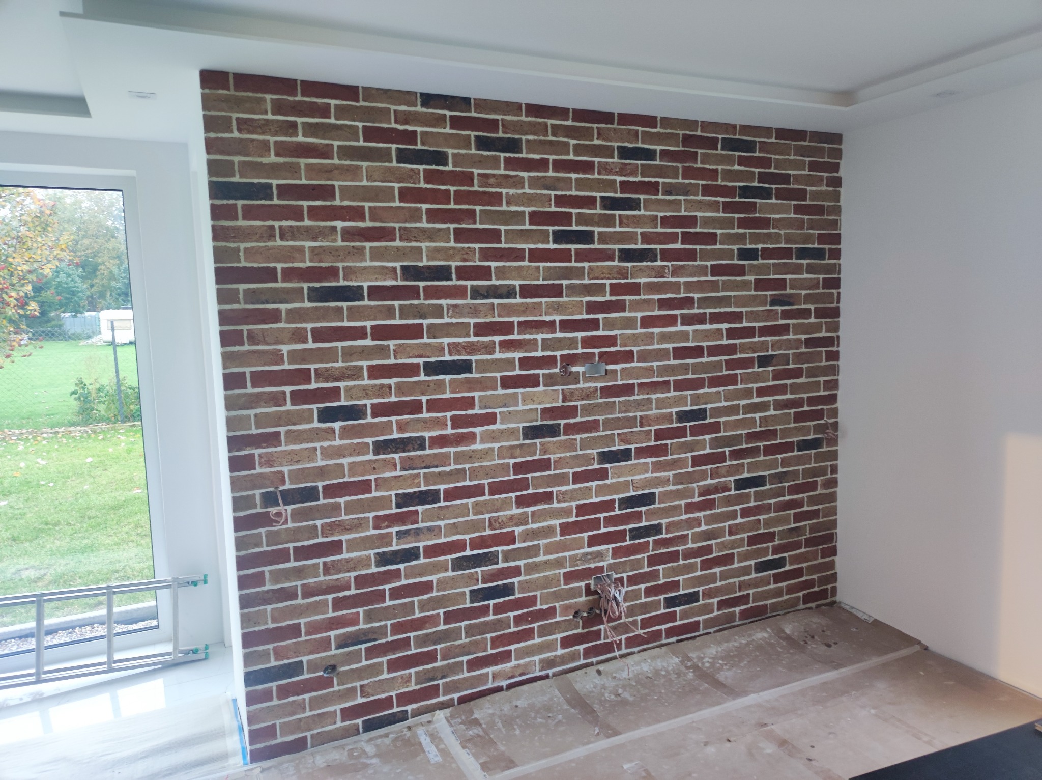 Ściana wykończona cegłą dekoracyjną w odcieniach czerwieni, brązu i czerni, z widocznymi przewodami elektrycznymi wystającymi ze ściany, fragment okna z drabiną opartą o parapet, zabezpieczona...