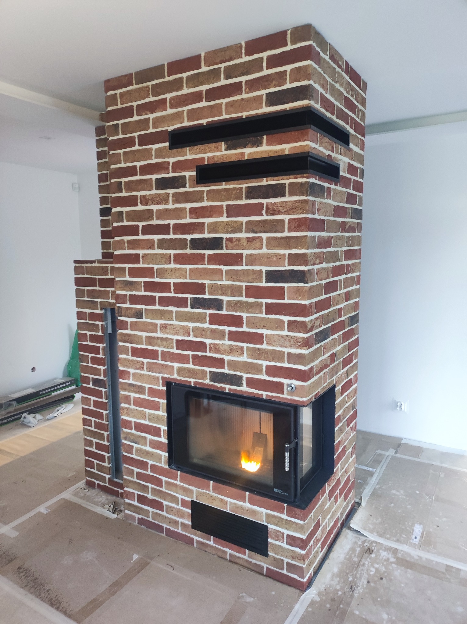 Nowoczesny kominek z cegły klinkierowej w odcieniach czerwieni i brązu, z czarnymi elementami dekoracyjnymi i paleniskiem z płomieniem, na podłodze zabezpieczonej kartonem.