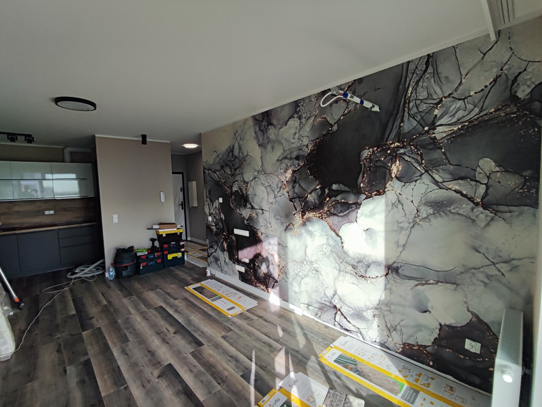 Pokój z fototapetą imitującą marmur w odcieniach szarości i złota, widoczne przygotowania do montażu z rozłożonymi opakowaniami paneli podłogowych Vilo i narzędziami.